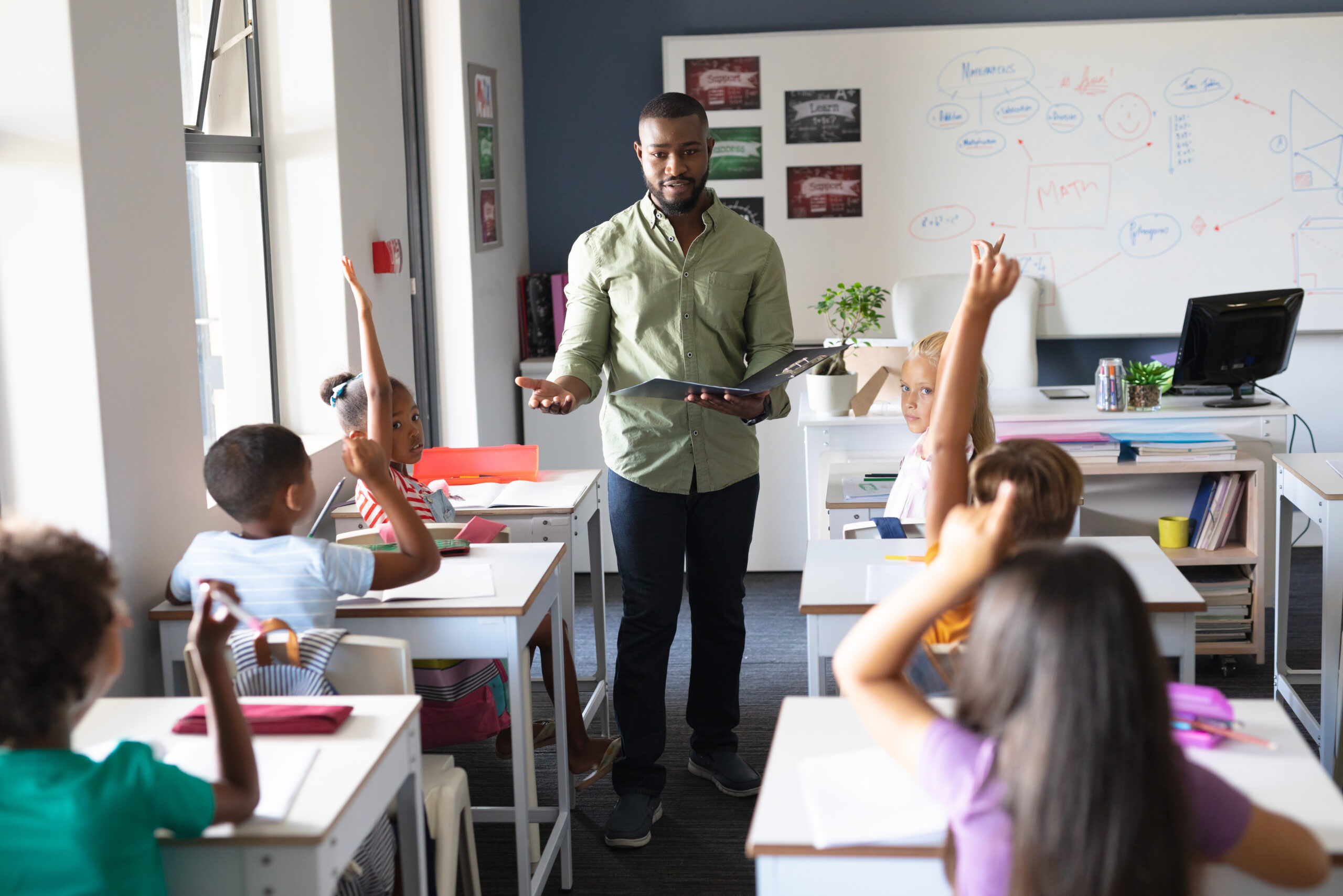 African american young male teacher with file standing by multiracial students with hand raised