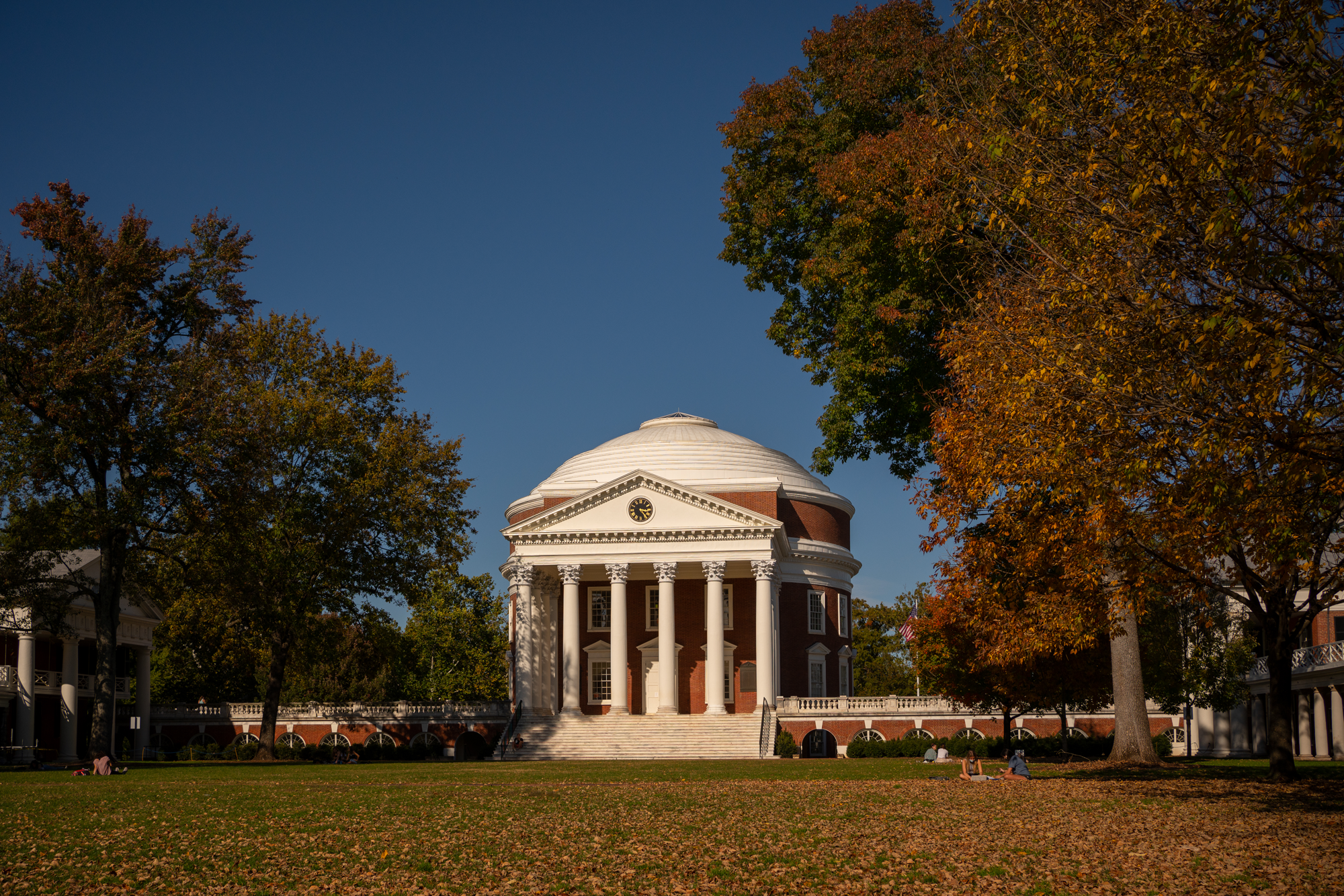 Domed university rotunda with columned portico, framed by autumn trees, students relaxing on lawn