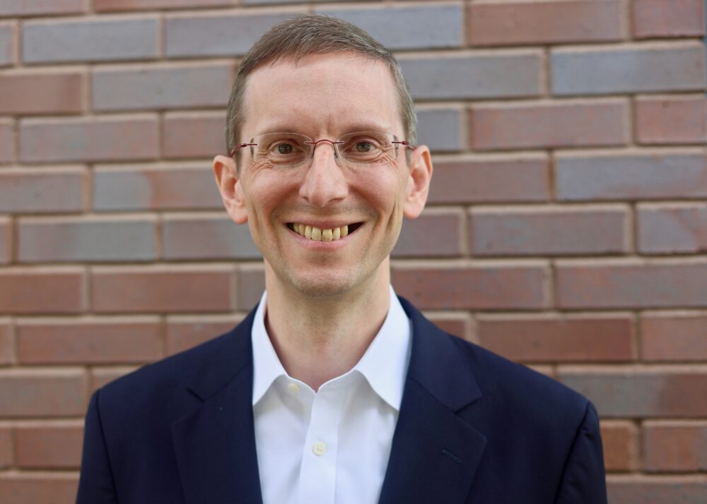 Smiling man with glasses wearing a white shirt and navy blazer, standing in front of a brick wall